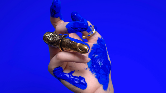 photo of an extended hand with a dark royal blue long sleeve, photographed in front of a Yves Klein blue background, with ultramarine blue paint splashed all over it and a golden all-finger ring on her finger
