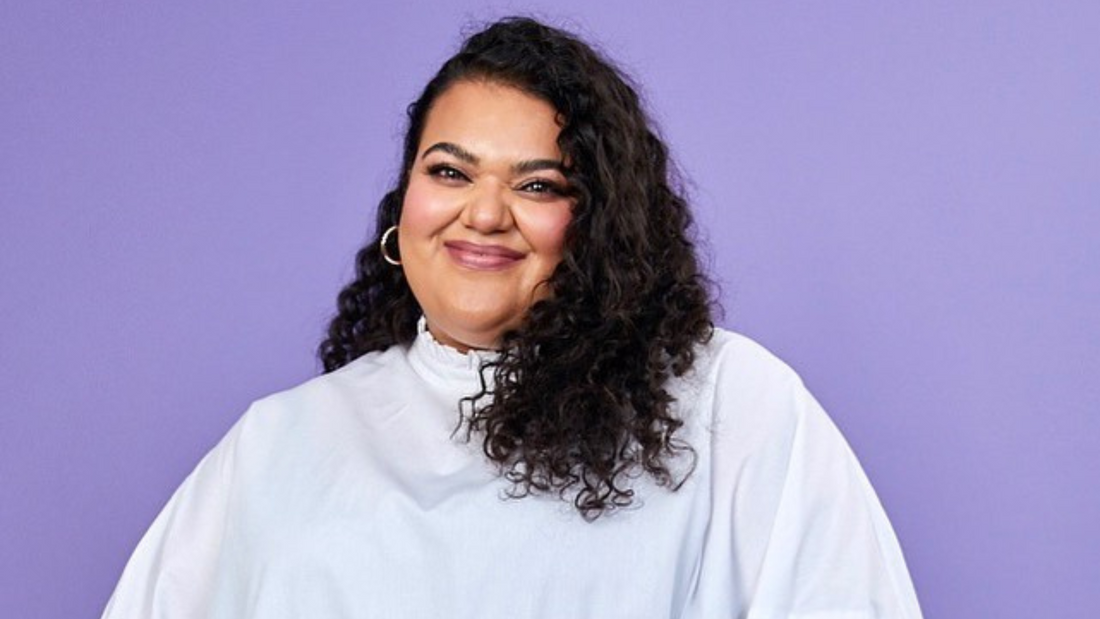Photo of a smiling woman of Indian heritage, with black curly hair with below shoulder length and wide white shirt, photographed in front of a purple background