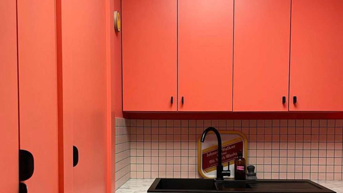 close up of a kitchen cupboard area with three visible kitchen cupboard painted in a coral orange colour, with added matt black handles, supported by a small area of square tiles and a matt black sink, YesColours