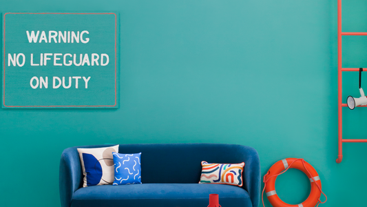 photo of a navy blue teal sofa photographed in the middle of a teal wall, with an orange ladder and life ring on the right and a sign saying 'warning no lifeguard on duty' on the left