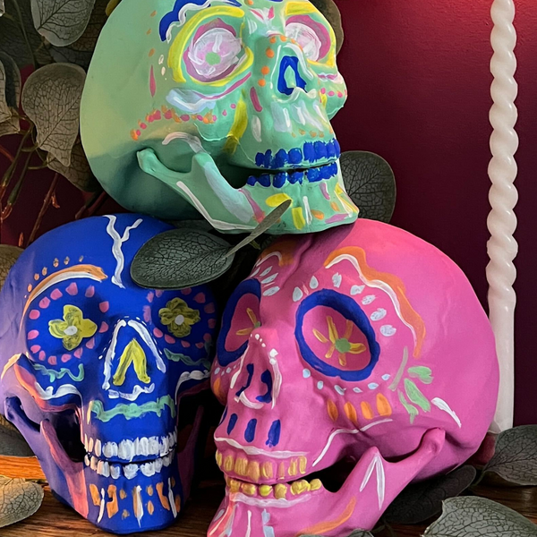 image of three artificial skulls in blue, green and pink and with wavy lines and dots decorated on them, stacked on top of each other next to a tall candle, small tea candle, leafy plant, all photographed in front of a magenta coloured purple pink wall
