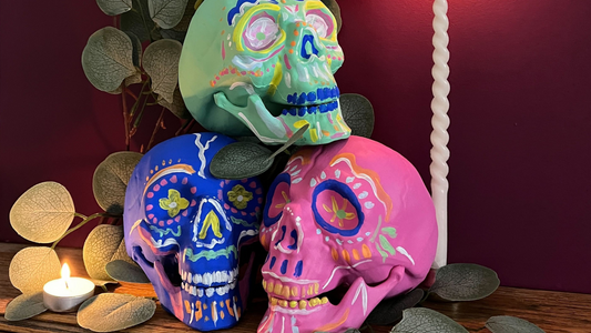 image of three artificial skulls in blue, green and pink and with wavy lines and dots decorated on them, stacked on top of each other next to a tall candle, small tea candle, leafy plant, all photographed in front of a magenta coloured purple pink wall