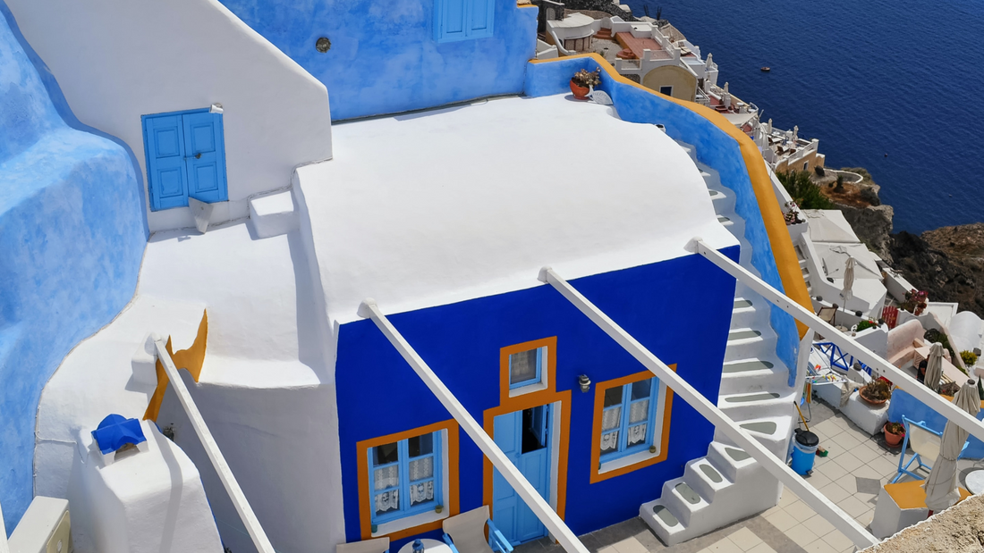 Photo of an island architecture consisting in a bright ultramarine blue painted house with orange window frames and white rooftop, surrounded by a bright blue stone wall, overlooking a blue sea to the right