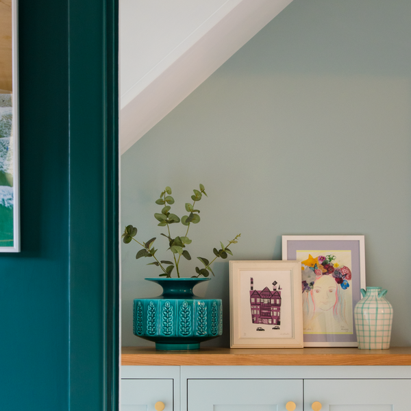 close up of a living room wall and a narrow hallway in dark and light green decorated with a nature-inspired sea painting, small artworks and a leafy plant in a large teal plant pot placed on a wooden light green sideboard