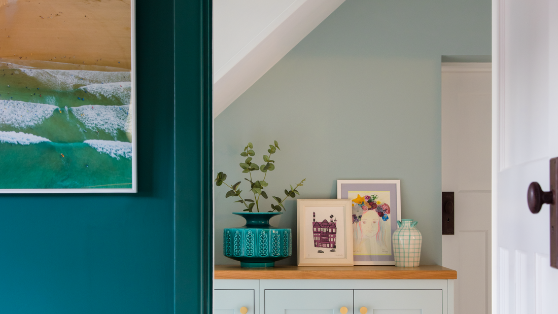 close up of a living room wall and a narrow hallway in dark and light green decorated with a nature-inspired sea painting, small artworks and a leafy plant in a large teal plant pot placed on a wooden light green sideboard