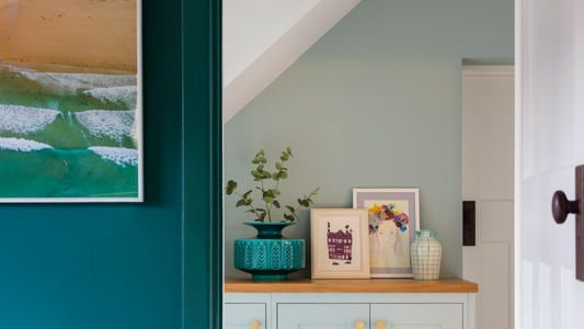 close up of a living room wall and a narrow hallway in dark and light green decorated with a nature-inspired sea painting, small artworks and a leafy plant in a large teal plant pot placed on a wooden light green sideboard