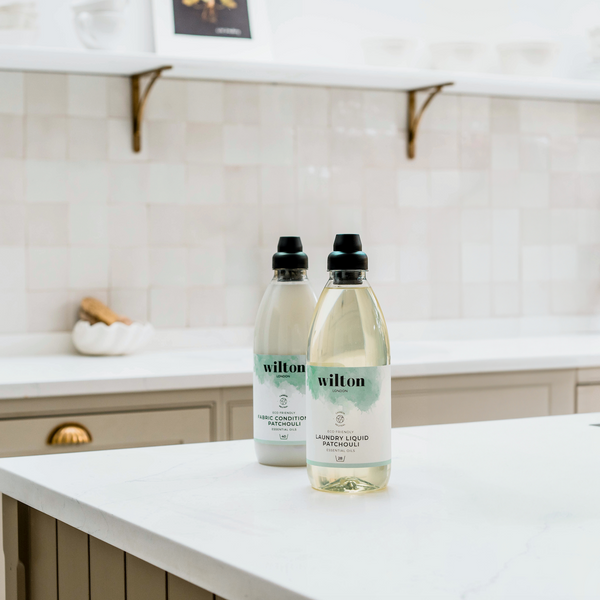 photo of a spacious contemporary kitchen with light beige square tiles, light brown coloured cupboards and a kitchen island with a white countertop, with two plastic bottles filled with liquid placed on top of it
