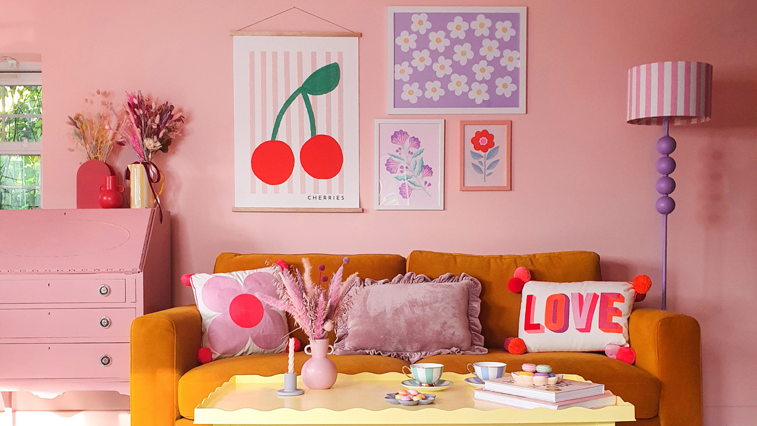 photograph of a mustard yellow sofa placed in the middle of a peachy pink coloured room with a dusky pink vintage dresser to the left and tall striped lamp on the right, surrounded by colourful artworks in lilac, red, pink and blue