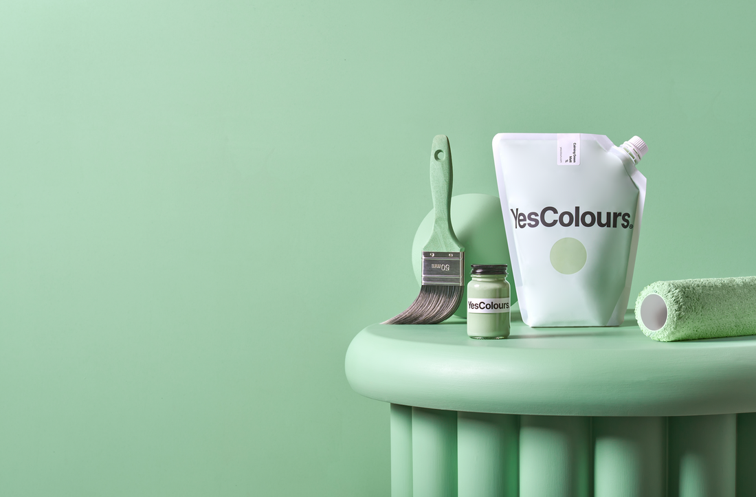 photograph of paint brush placed vertically, small glass jar full of green paint, green ball, pouch and green-coloured paint roller, placed on a green table and photographed on a green background