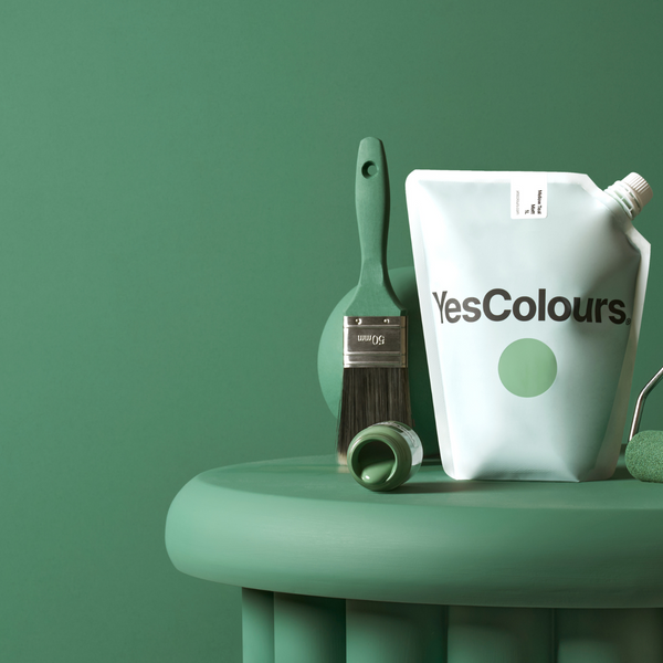 photo of a white pouch with a logo that reads YesColours, surrounded by a paint brush and roller and green coloured ball, all placed on a green painted ribbed side table and photographed in front of a forest green wall