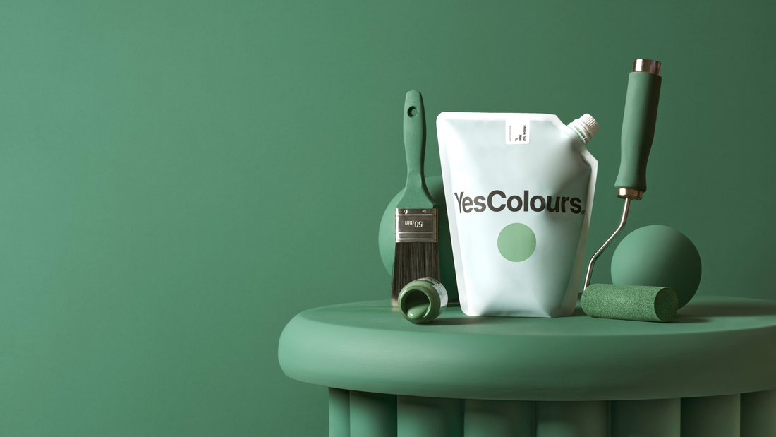photo of a white pouch with a logo that reads YesColours, surrounded by a paint brush and roller and green coloured ball, all placed on a green painted ribbed side table and photographed in front of a forest green wall