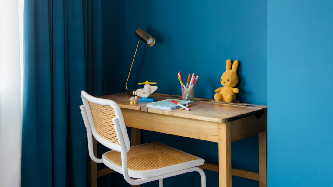 photo of a children's bedroom cover with a vintage wooden desk, white and rattan chair as well as a plush bunny toy, crayons and a table lamp placed on top of it, surrounded by a dark teal colour on the walls and a peachy pink colour on the ceiling