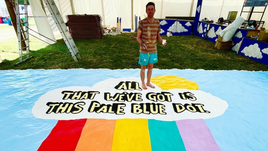 a man dressed in orange stripe pattern t-shirt and aqua coloured summer shorts, stepping on an artwork in a pale blue colour with rainbow stripes in red, orange, yellow, teal and lilac and text that reads all that we've got is this pale blue dot