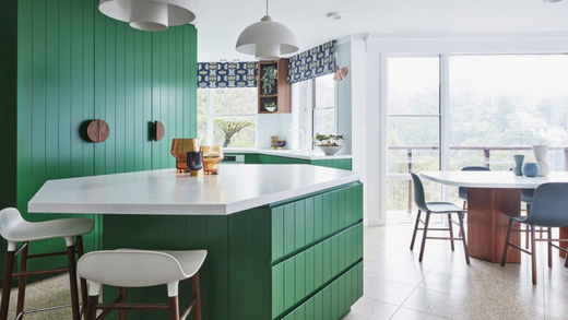 forest green painted kitchen cabinets and kitchen island, surrounded by tall wicker chairs in white, overlooking an airy dining room with table and chairs