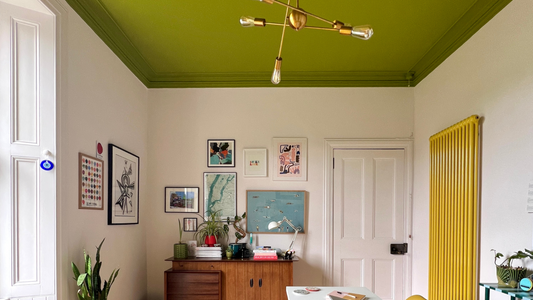 photograph of a home office with neutral walls and a big window on the left with natural light coming in, an olive green ceiling, bright yellow radiator on the right, dark wooden floor, gallery wall and a plain white desk placed on the right side