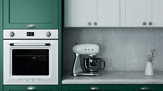 a minimalist kitchen with dark green painted cupboards, integrated oven on the left side, white kitchen cupboards in the middle and a concrete blacktop and countertop with a white Smeg mixer and small vase placed on top