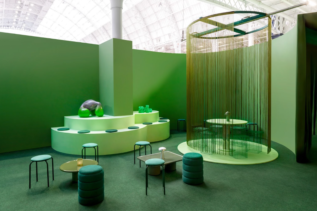 A modern interior seating area with green walls and carpet, featuring tiered light green platforms, small round tables, dark green stools, and a tall cylindrical glass enclosure with hanging rods.