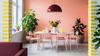 A dining room scene with a white rectangular table and four pink chairs, set against a peach-pink wall. Yellow-and-white striped columns frame the scene, with potted green plants on either side.