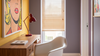 A modern home office corner featuring a wooden desk with a red adjustable desk lamp, a white molded chair, a window with beige blinds, and a large pop-art style framed print on a yellow wall.
