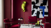 A modern living room scene with deep red armchairs, a matching red sofa, a yellow sculptural floor lamp, and a large abstract black, white, and blue wall art piece against a dark red wall.