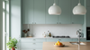 A modern kitchen interior with pale sage green cabinets, white countertops and backsplash, two white pendant lights, a wooden island, and a bowl of oranges on the counter.