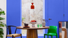 A styled interior scene with a round wooden dining table in the center, flanked by a black chair with a beige seat and a bright green chair. A red pendant light hangs above the table, which holds a bowl of fruit, a patterned vase, and green glassware.